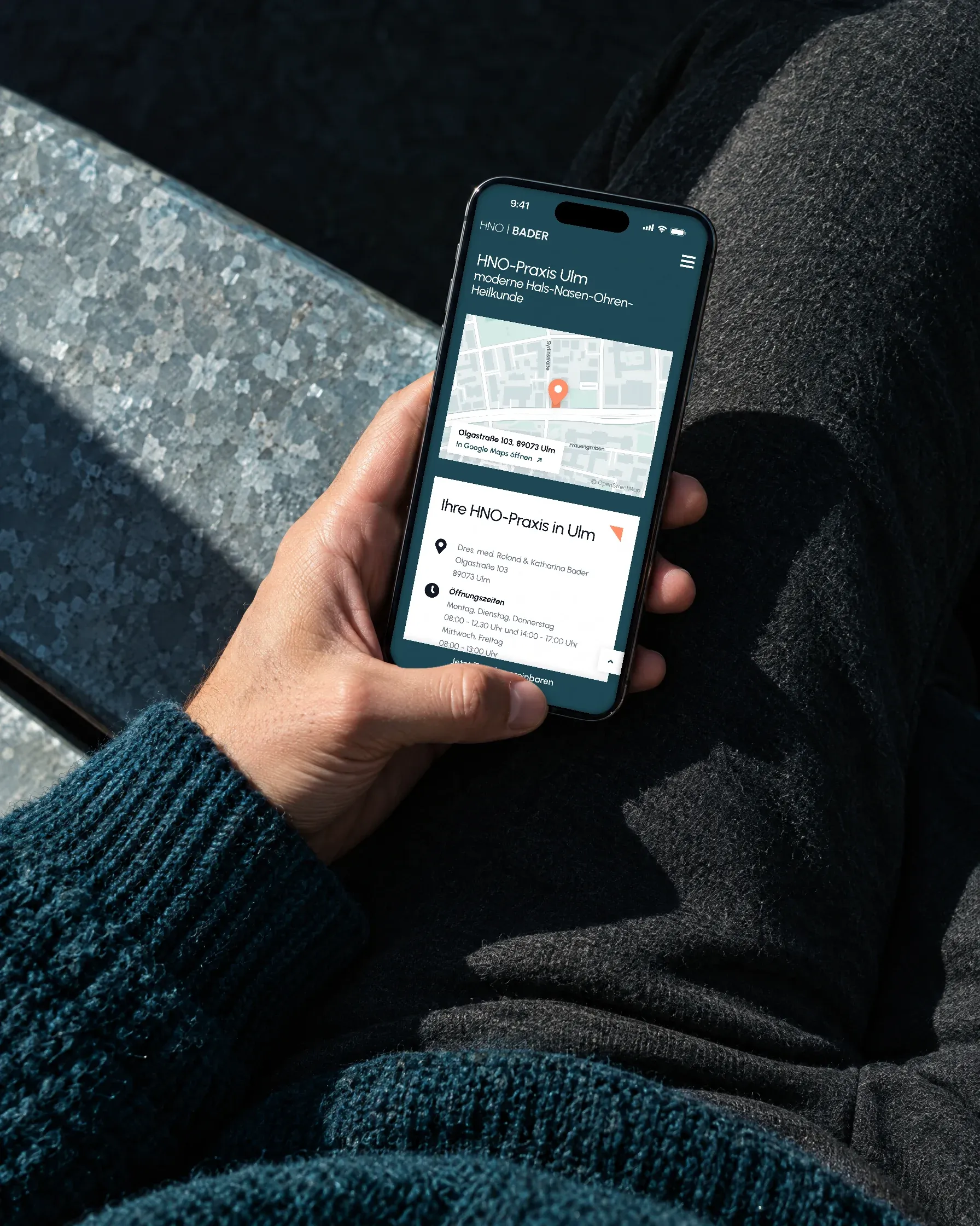 Eine Hand hält ein Handy mit Lageplan und Öffnungszeiten der HNO-Praxis Bader, die Person sitzt im Sonnenlicht auf einer Metallbank