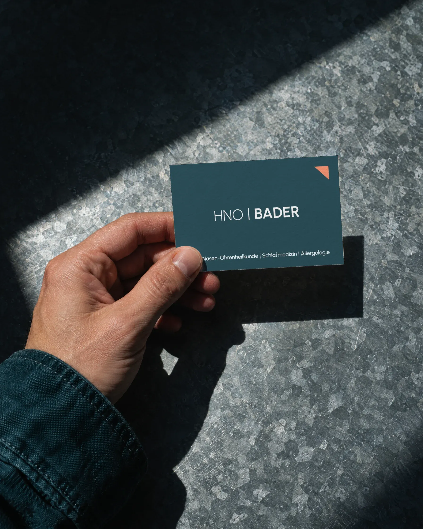 Eine Hand hält eine Terminkarte der HNO-Praxis Bader mit der Vorderseite nach oben über verzinktes Blech