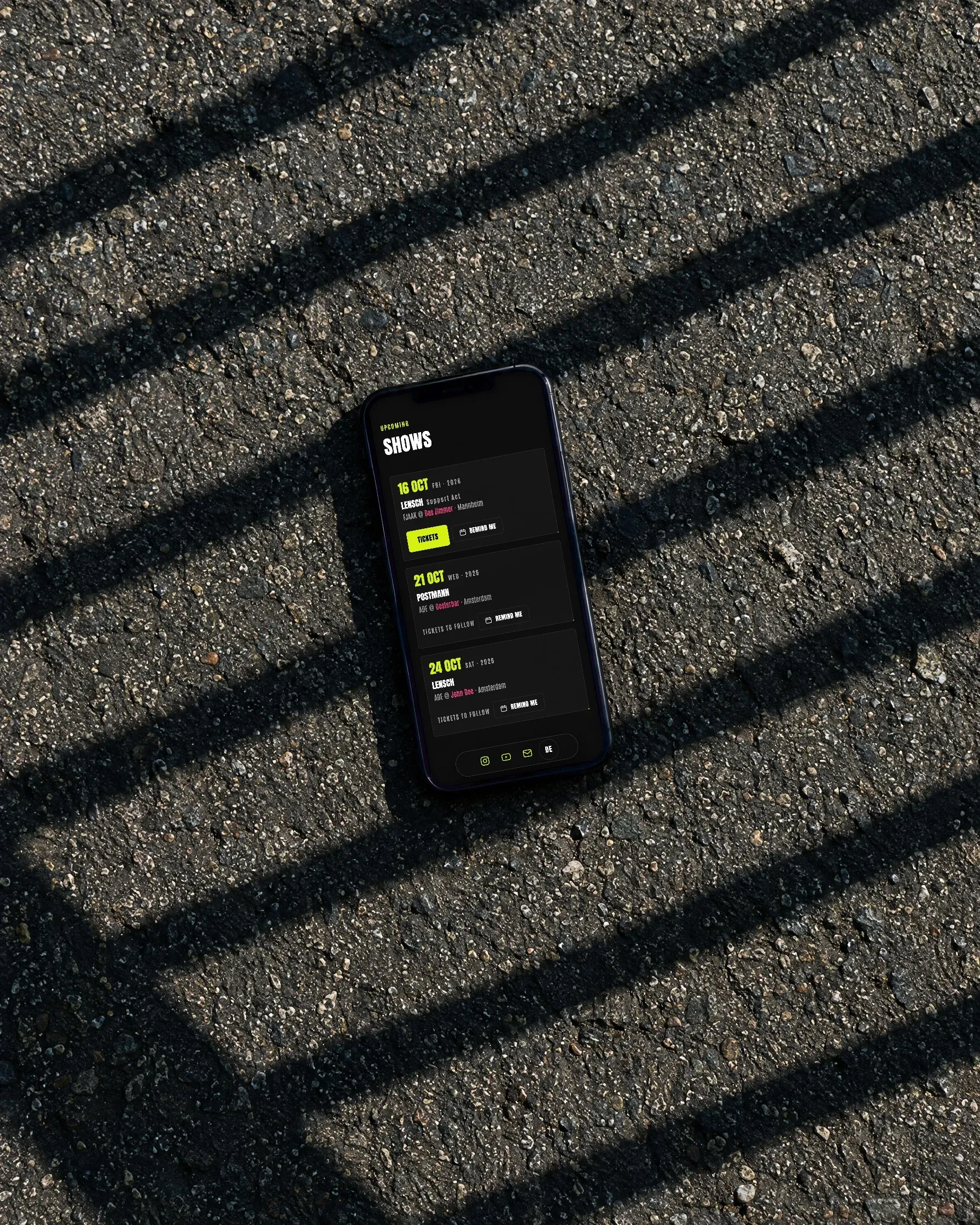 Ein Handy mit dem Showkalender von Joscot liegt auf Asphalt im Schatten eines Geländers