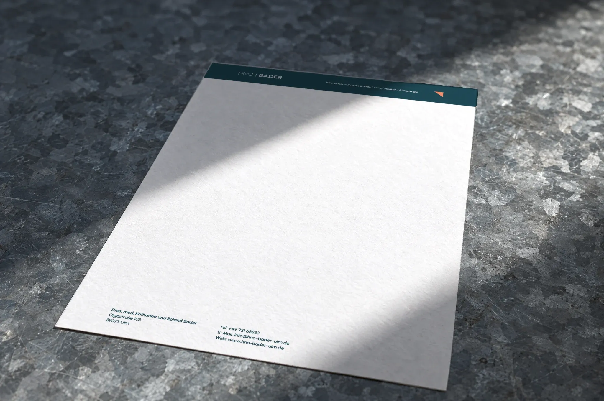 Ein Bogen Briefpapier der HNO-Praxis Bader liegt auf verzinktem Blech, ein Streifen Sonnenlicht fällt darüber