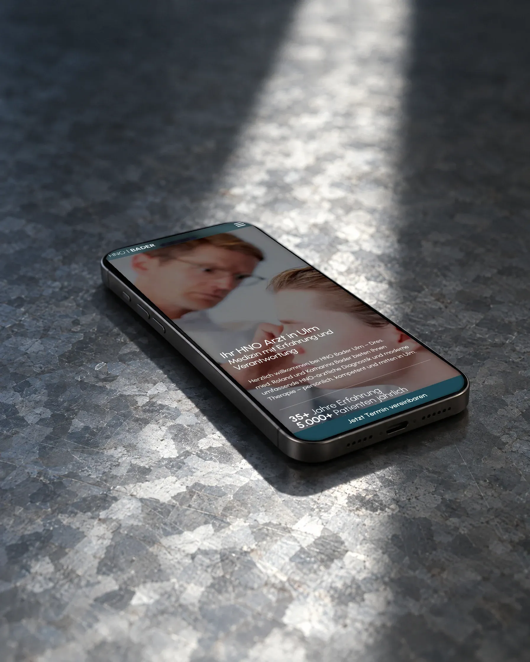 Ein Handy mit der Startseite der HNO-Praxis Bader liegt auf verzinktem Blech, dahinter ein Streifen Sonnenlicht
