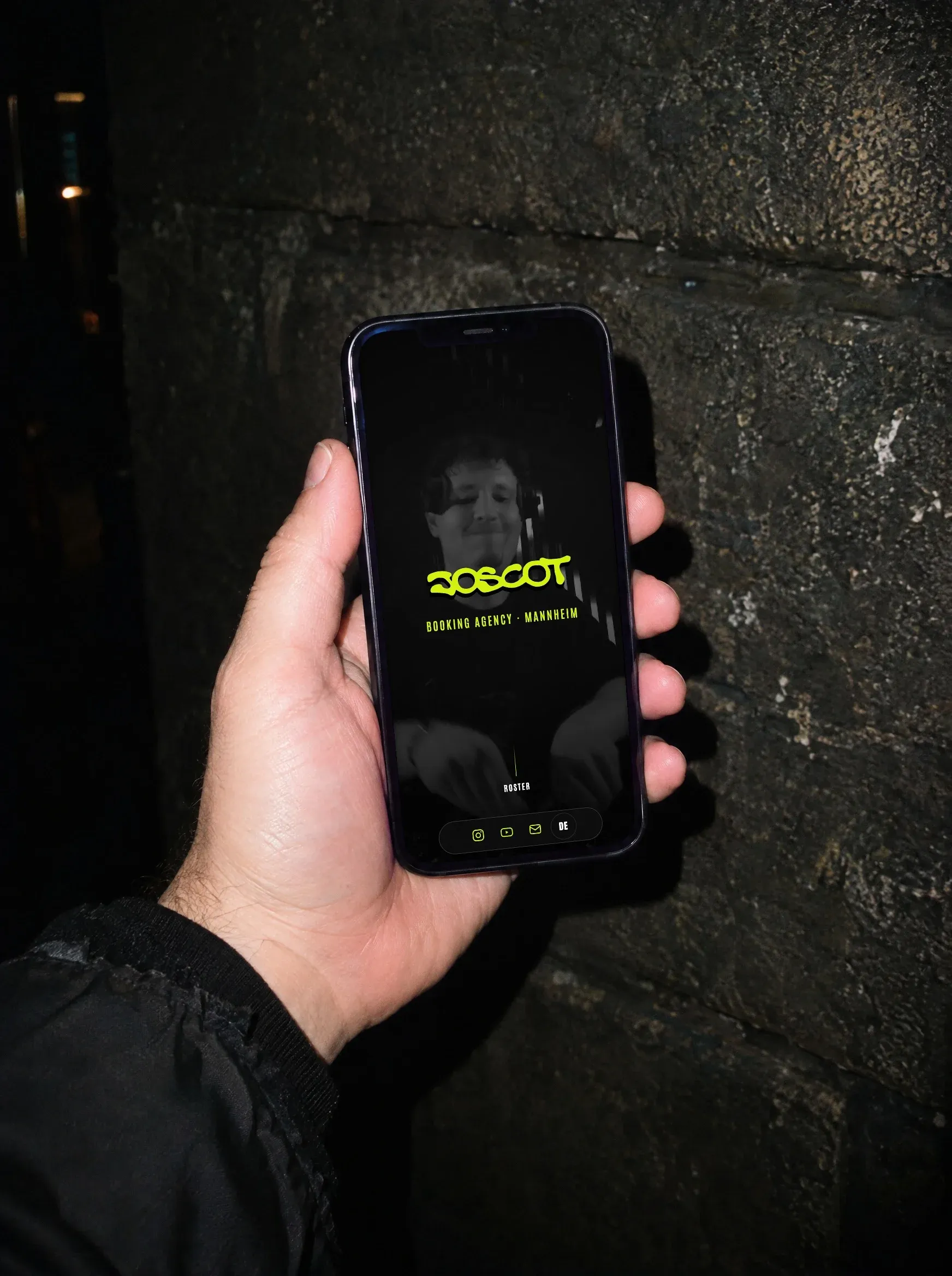 Eine Hand hält nachts ein Handy mit der Startseite von Joscot vor eine Betonwand
