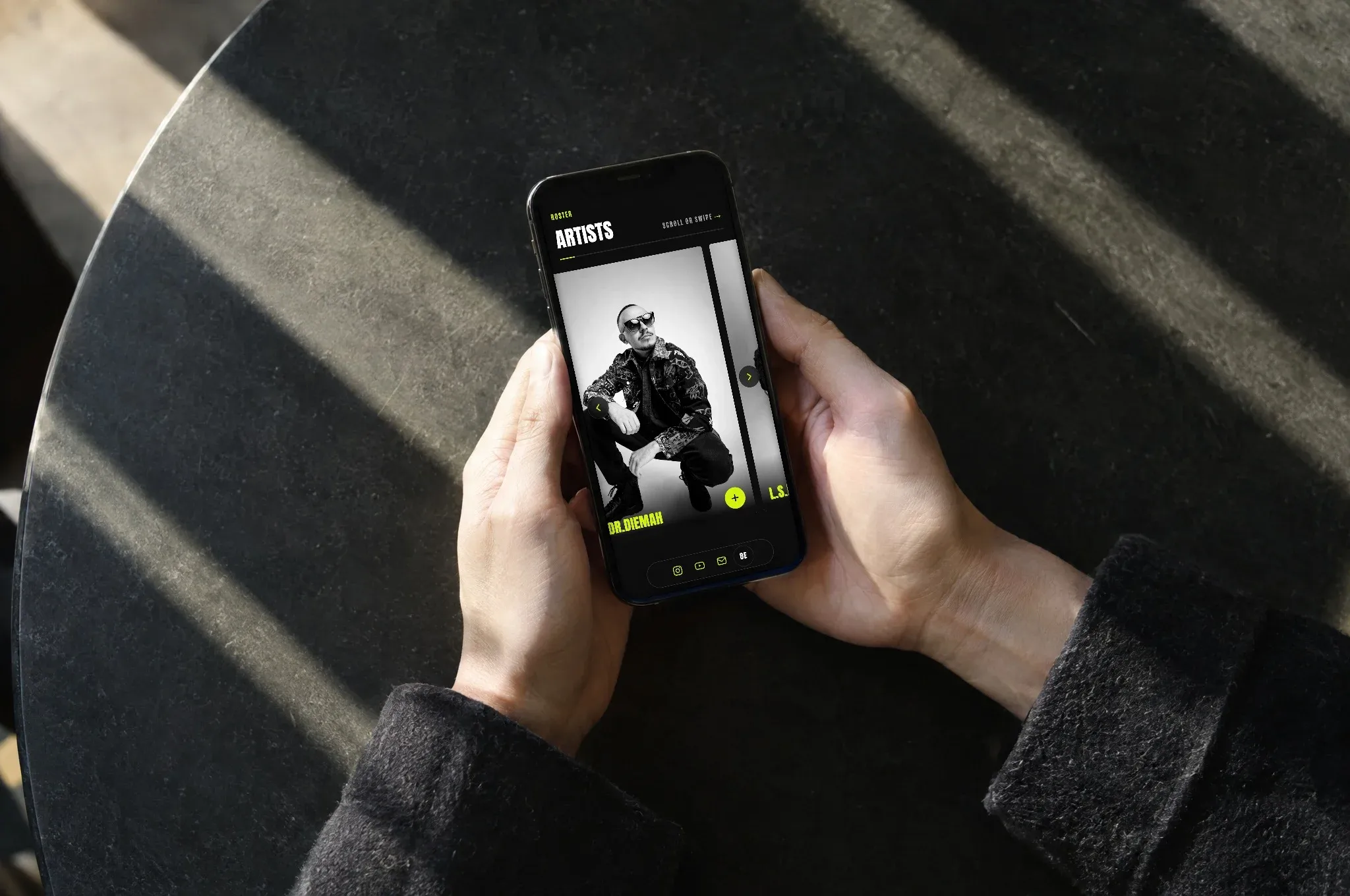 Zwei Hände halten ein Handy mit der Artist-Übersicht von Joscot über einem dunklen Tisch