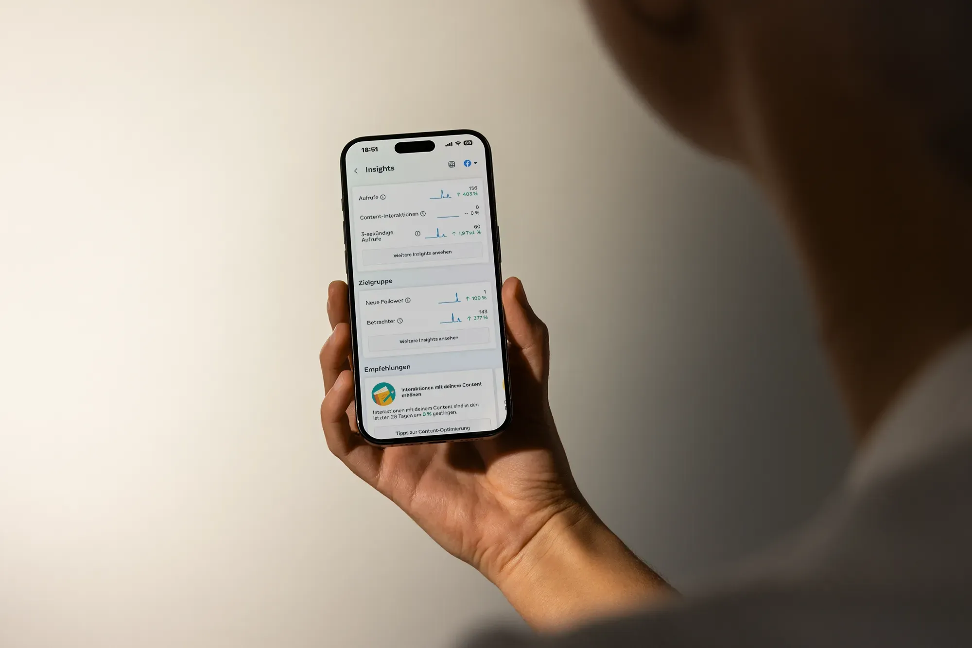 Eine Hand hält ein Handy vor einer hellen Wand, auf dem Bildschirm die Statistik einer Facebook-Seite