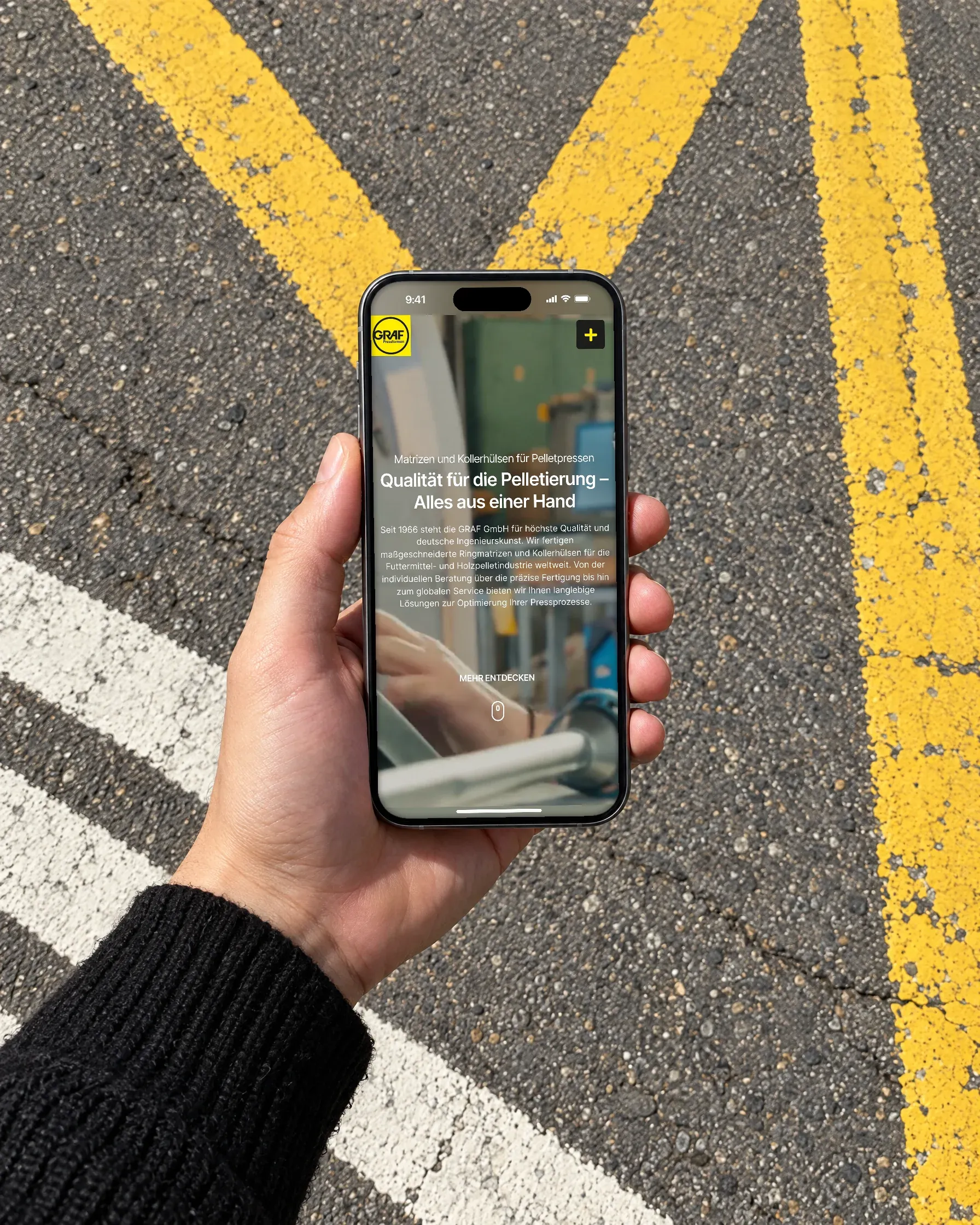 Eine Hand im schwarzen Pullover hält ein Handy mit der deutschen Startseite der Graf GmbH über Asphalt mit gelben Fahrbahnmarkierungen