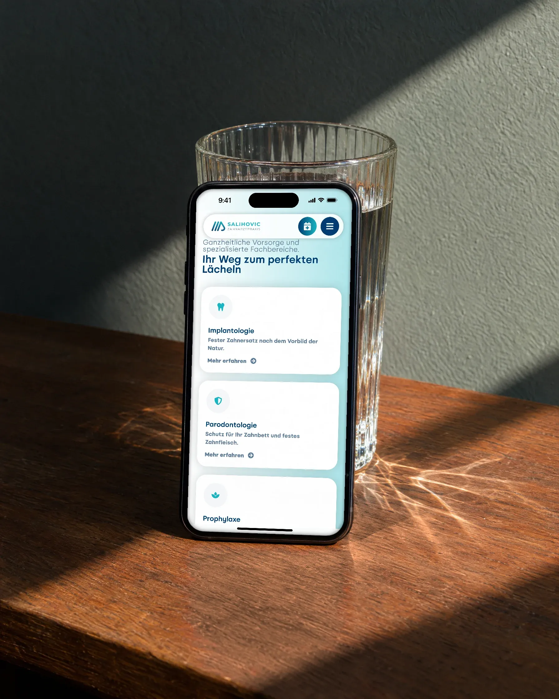 Ein Handy mit den Leistungen der Zahnarztpraxis Salihovic lehnt auf einem Holztisch an einem Wasserglas