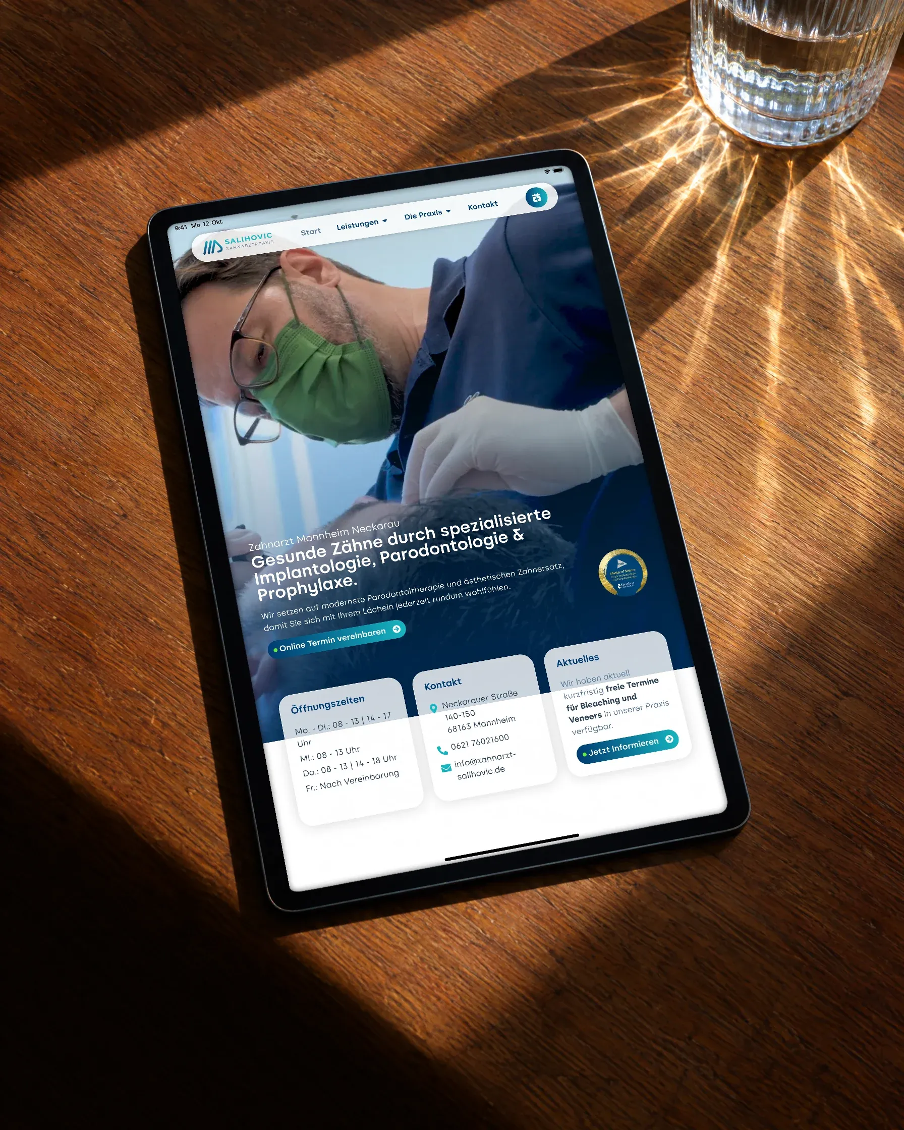 Ein Tablet mit der Startseite der Zahnarztpraxis Salihovic liegt auf einem Holztisch im Sonnenlicht, daneben ein Wasserglas mit Lichtfächer