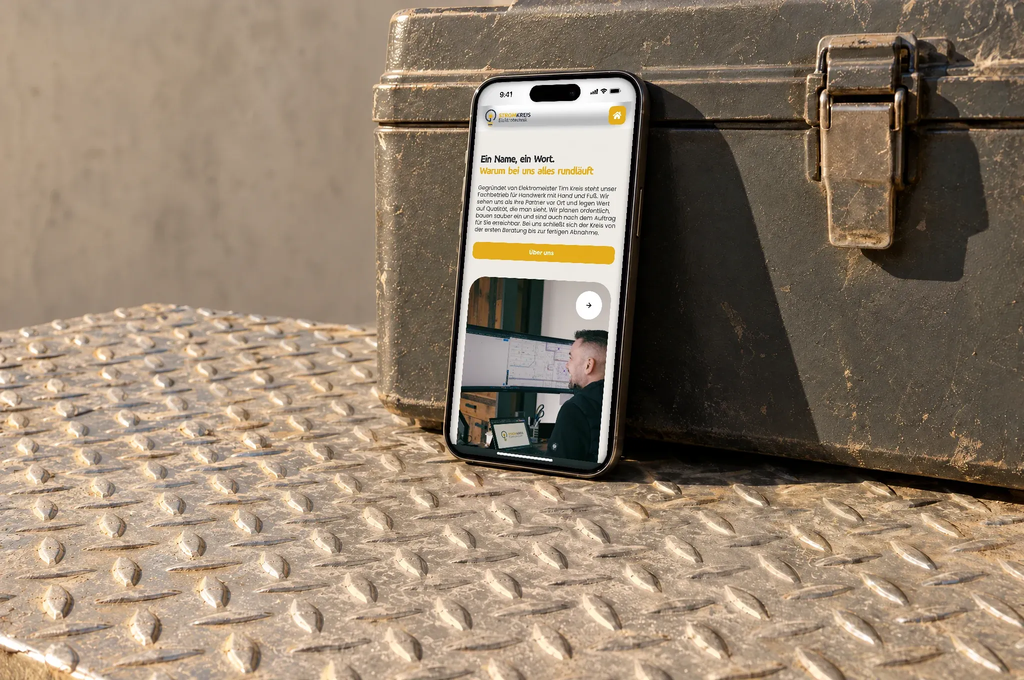 Ein Handy mit der Website von Stromkreis Elektrotechnik lehnt in der Sonne an einem Werkzeugkoffer auf Riffelblech