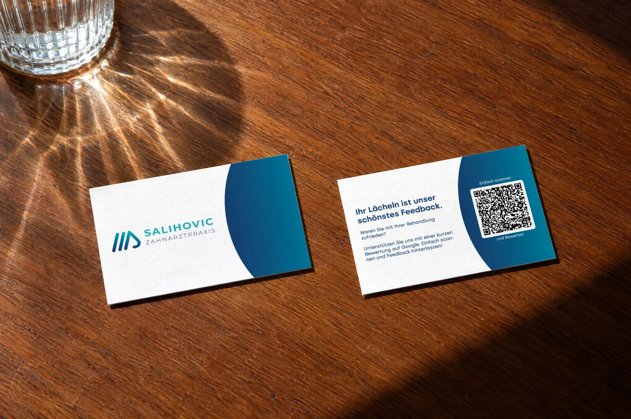 Zwei Karten der Zahnarztpraxis Salihovic liegen auf einem Holztisch neben einem Wasserglas, eine mit dem Logo, eine mit dem QR-Code für Bewertungen
