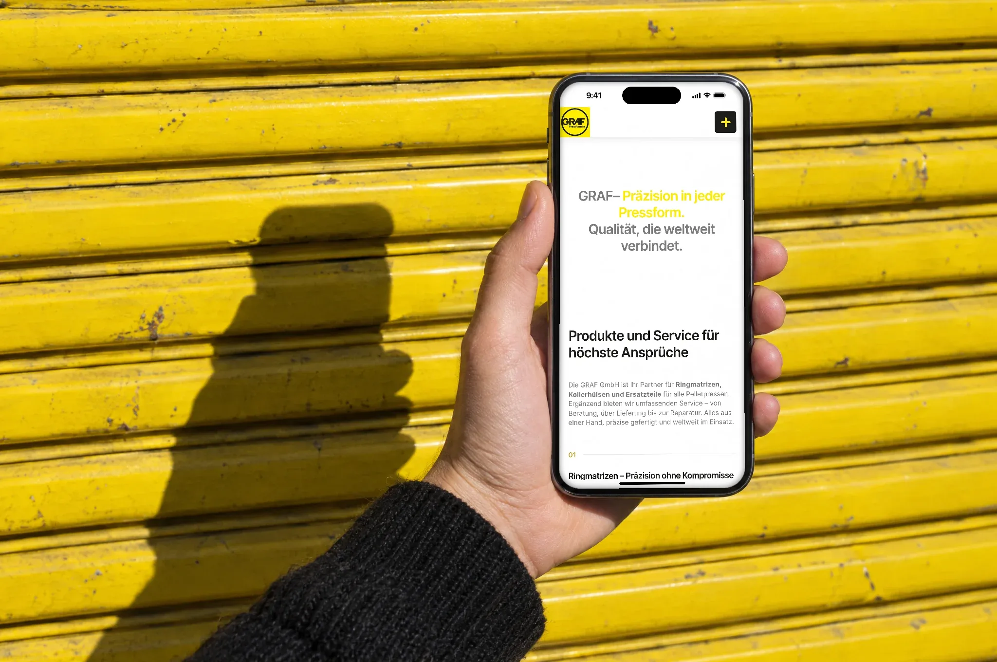 Eine Hand hält ein Handy mit der Graf-Website vor ein gelbes Rolltor in der Sonne
