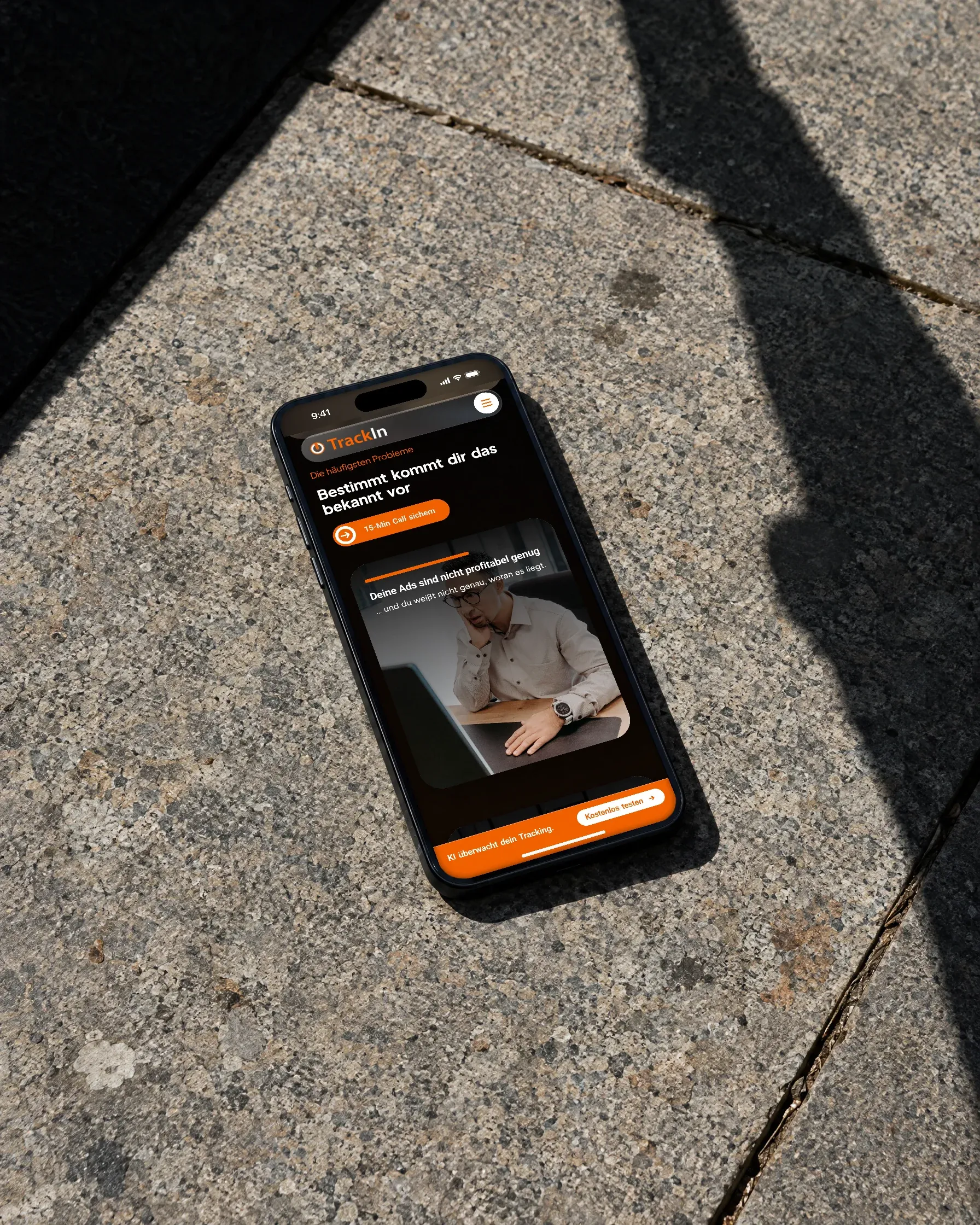 Ein Handy mit der Website von TrackIn liegt auf einer Granitplatte in der Sonne, daneben fällt der Schatten eines Menschen über das Pflaster