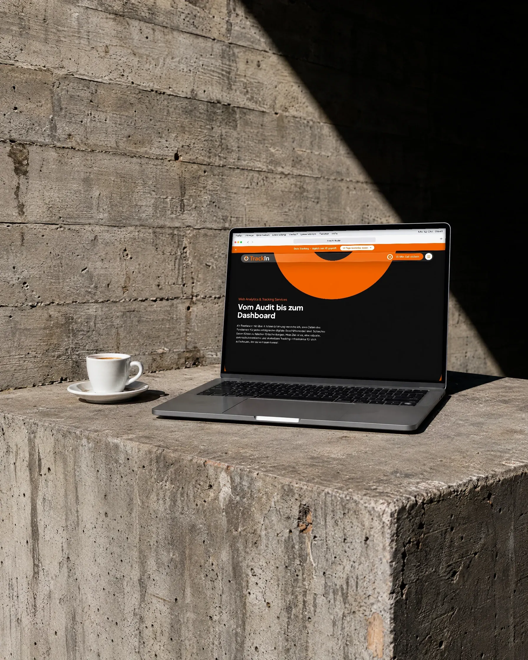 Ein Laptop mit der Leistungsseite von TrackIn steht auf einem Betonsockel, daneben eine Espressotasse