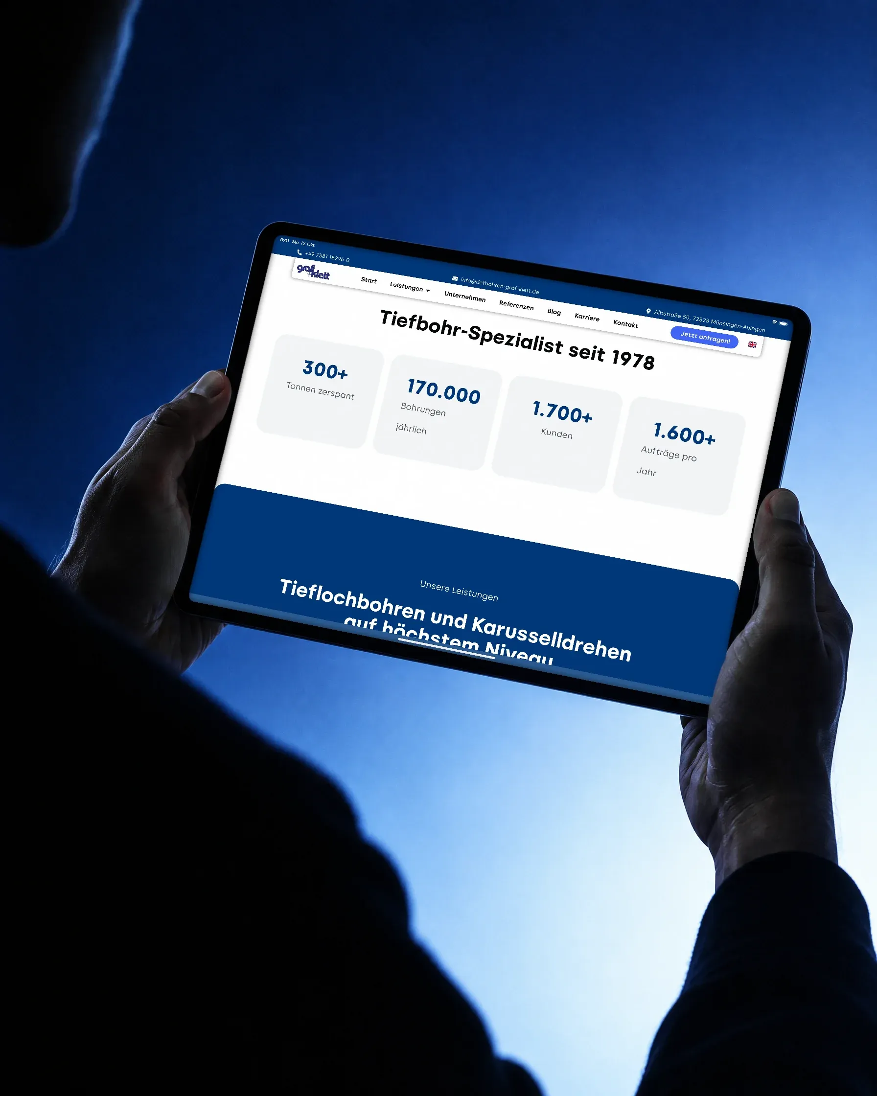 Über die Schulter gesehen: Zwei Hände halten vor blauem Verlauf ein Tablet quer, darauf die Zahlen des Betriebs auf der Website von Graf + Klett