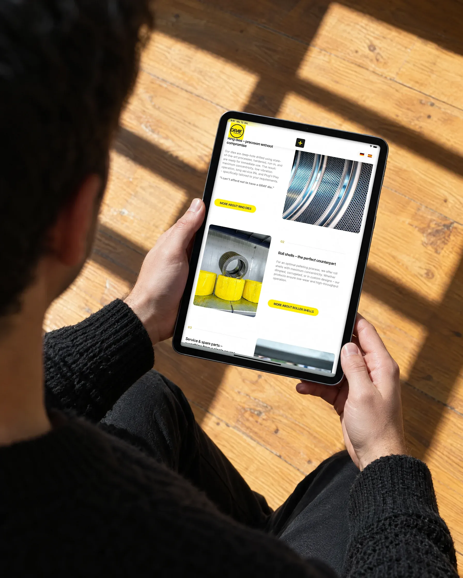 Blick über die Schulter: Zwei Hände halten ein Tablet mit der englischen Fassung der Graf-Website über einem sonnigen Holzboden