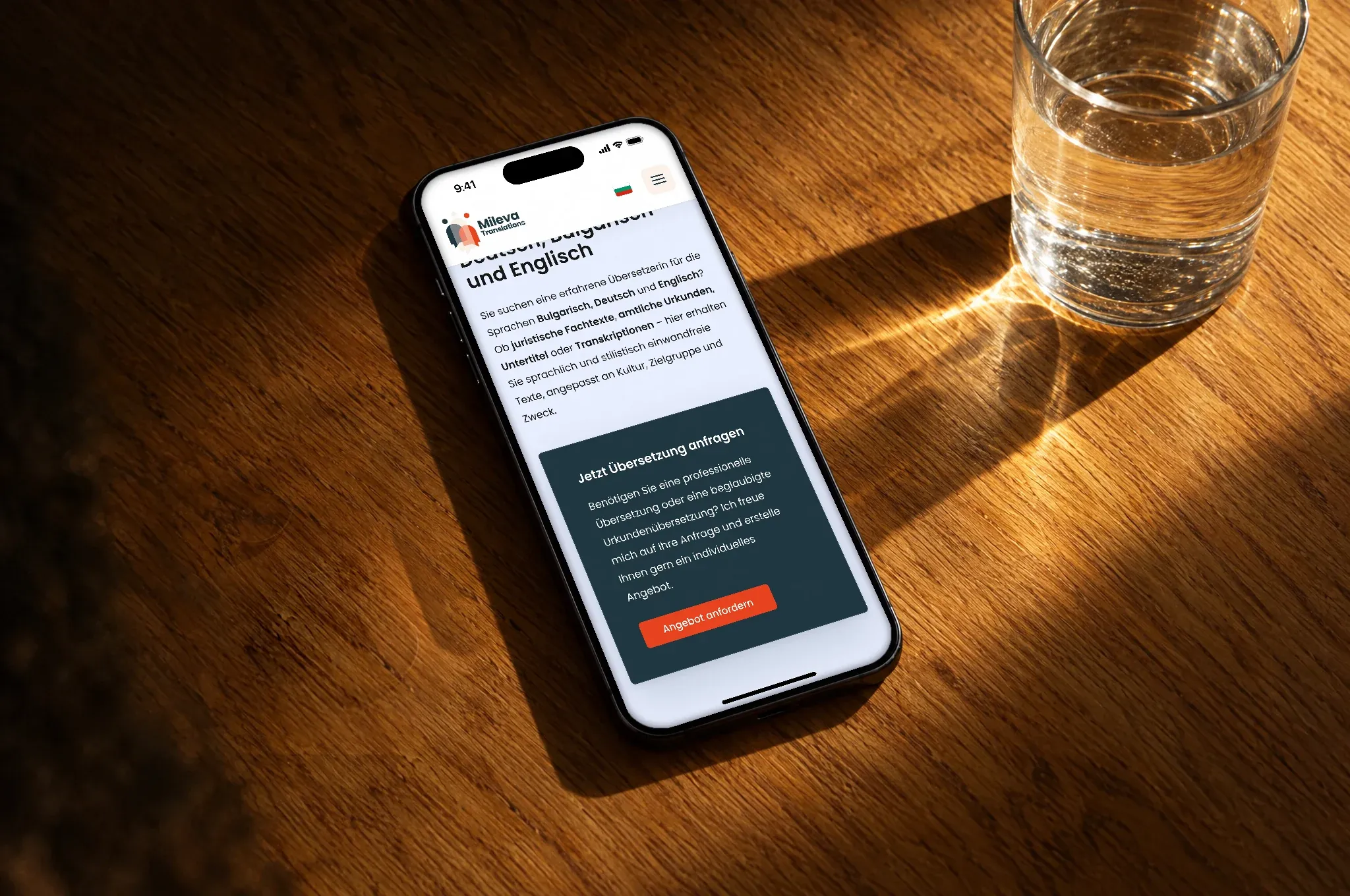 Ein Handy liegt auf einem Holztisch neben einem Glas Wasser und zeigt die Website von Mileva Translations in ihren Farben