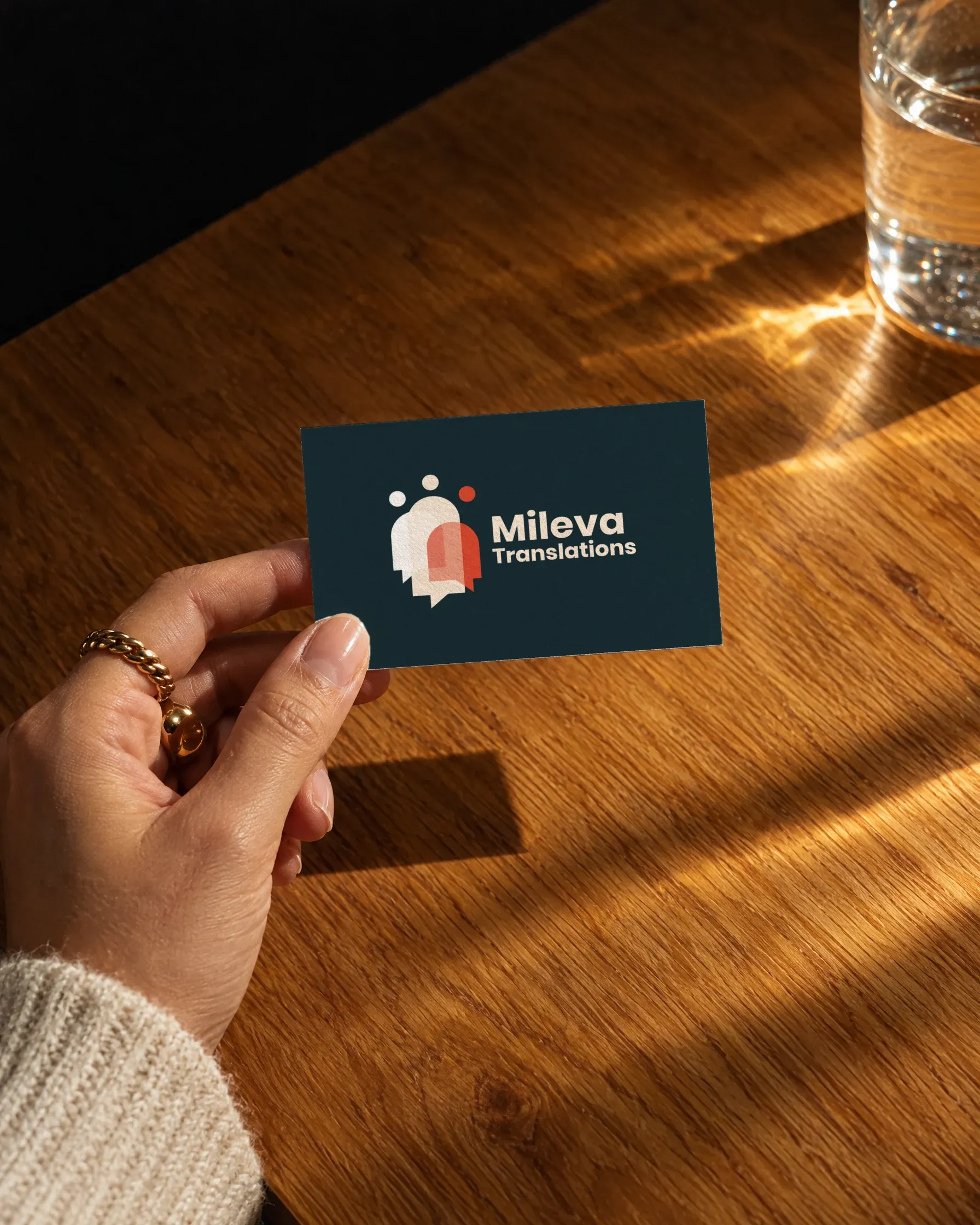 Eine Hand hält eine Visitenkarte von Mileva Translations mit dem Logo auf schieferblauem Grund über einen Holztisch