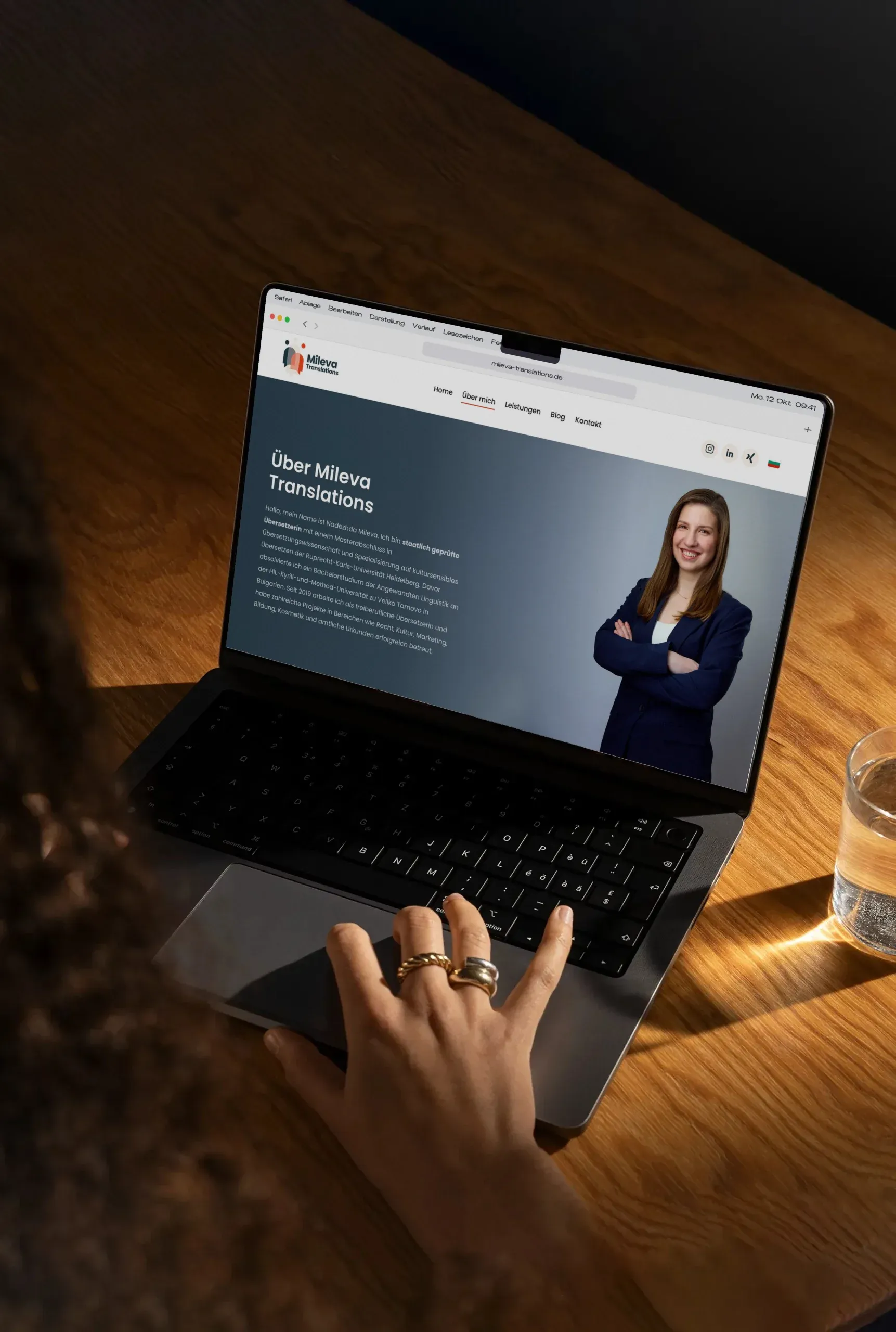 Blick über die Schulter auf einen Laptop mit der Seite „Über Mileva Translations", eine Hand mit Goldringen auf dem Trackpad