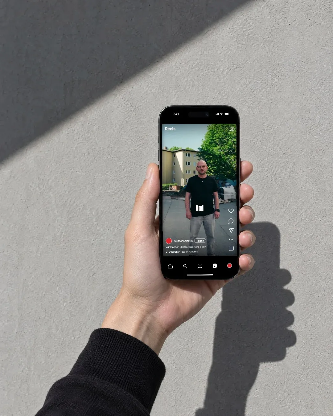 Eine Hand hält ein Handy vor eine helle Betonwand, darauf ein Recruiting-Reel der Deutschen Elektro