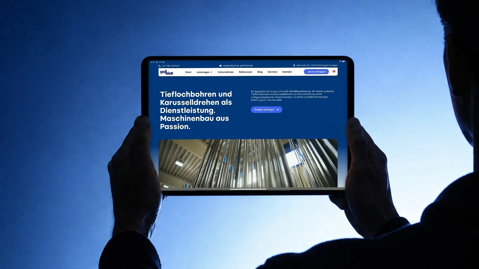 Eine Person hält als Silhouette vor einem blauen Farbverlauf ein Tablet hoch, auf dem die Website der Graf + Klett GmbH durchläuft