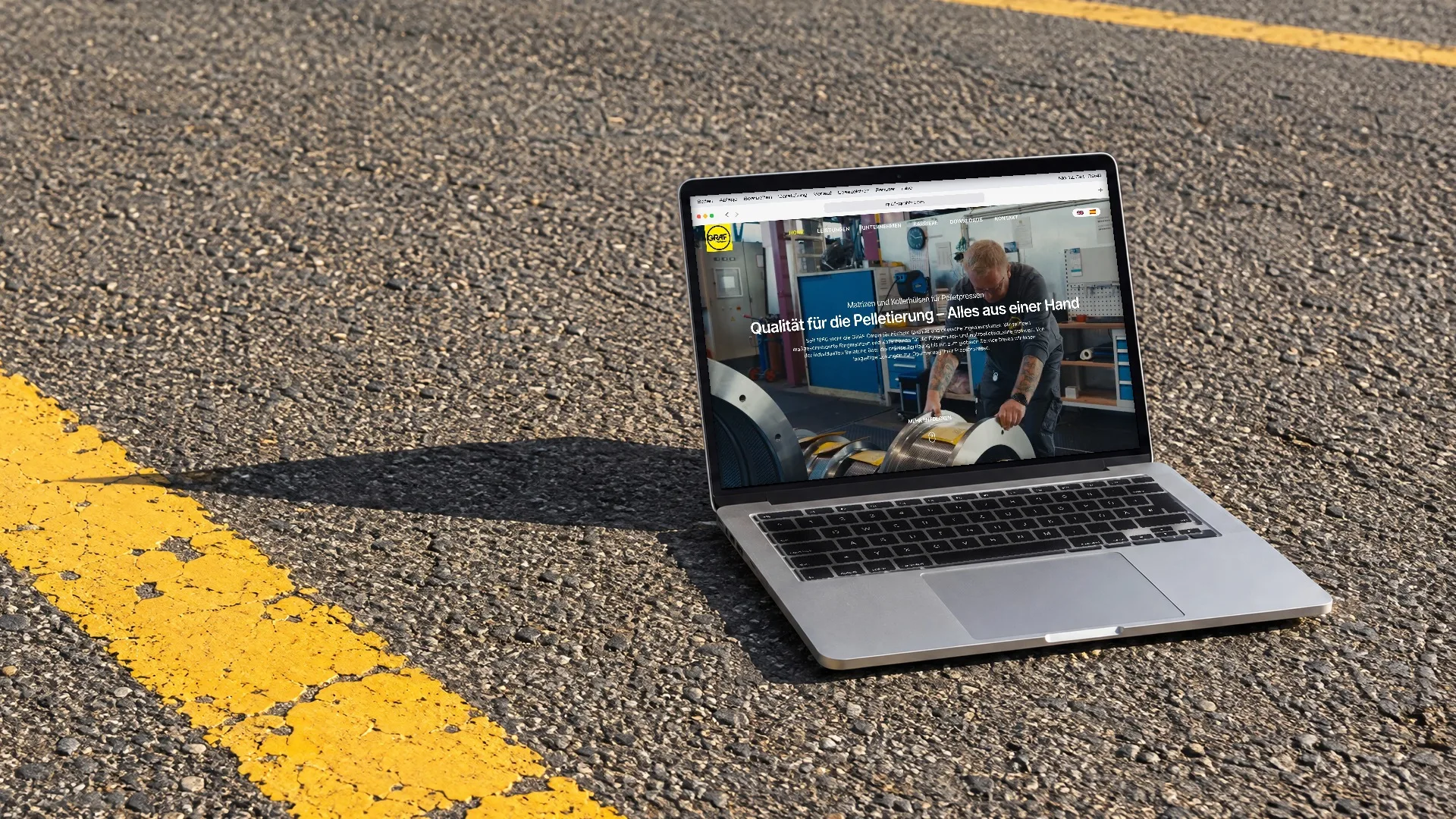 Ein Laptop steht in praller Sonne auf Asphalt neben einer gelben Fahrbahnmarkierung, auf dem Bildschirm läuft die Website der Graf GmbH durch