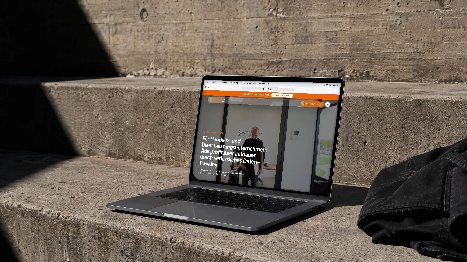 Ein Laptop steht auf einer Betonstufe in der Sonne, auf dem Bildschirm läuft die Website von TrackIn durch, daneben liegt eine schwarze Jacke